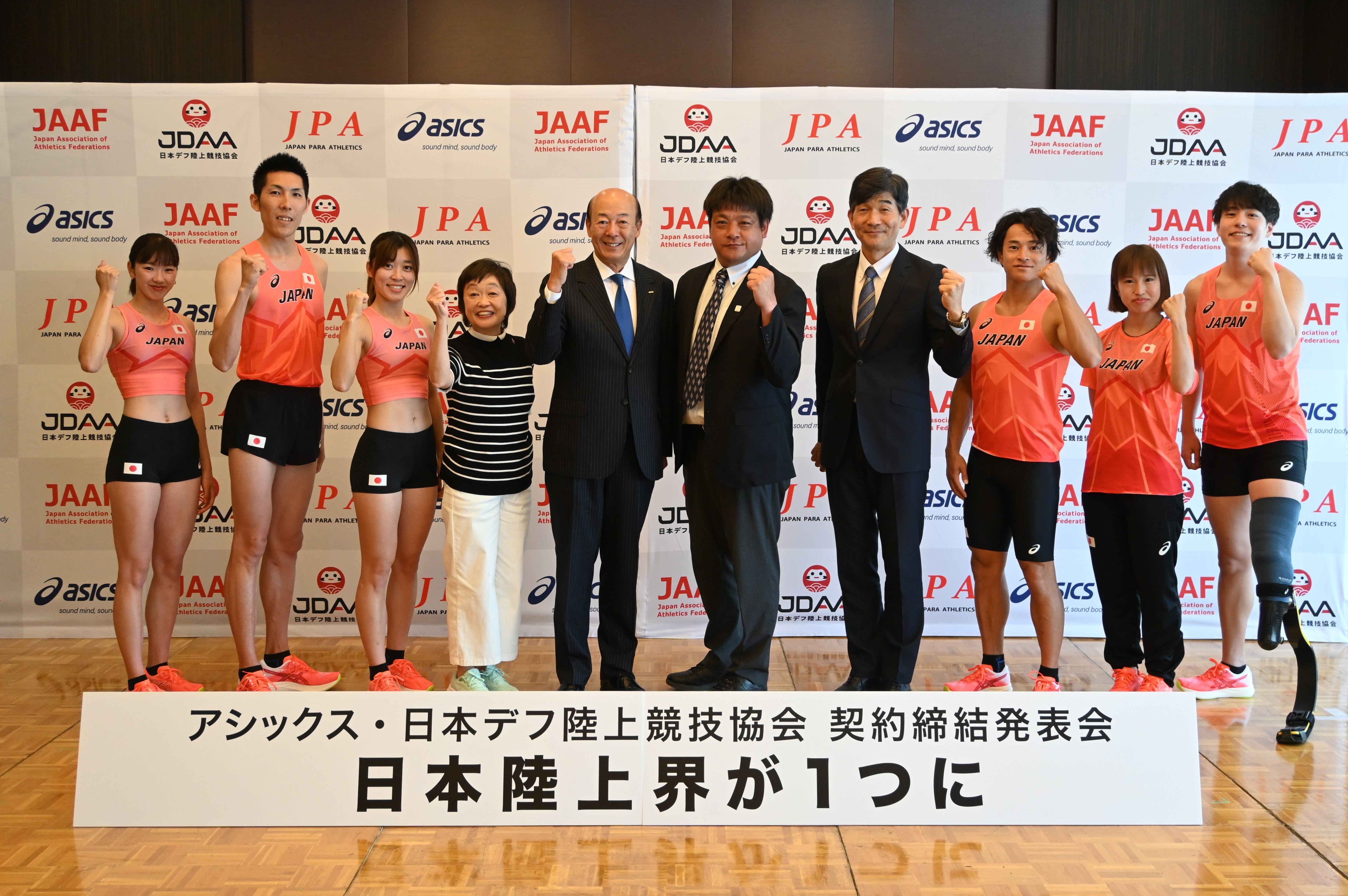 記者会見場で「日本陸上界がひとつに」の看板を前にガッツポーズをするアシックス廣田会長、増田明美さん、JDAA役員とアスリート、JPA役員とアスリート、計10名が並んだ写真